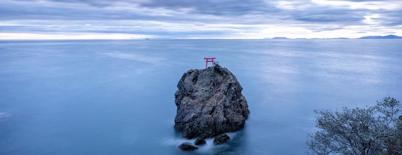  rundlicher Falesen liegt einsam im hellblauen, ruhigen Meer, darauf ein rotes japanisches Shinto-Tor
